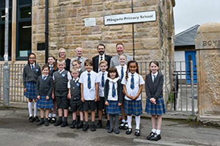 Group of adults and school pupils standing outside blond sandstone school building with a sign which reads Milngavie Primary School. 