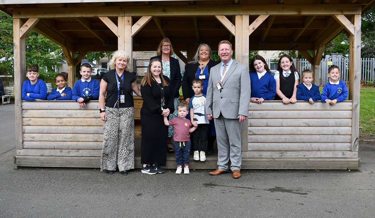 Children and adults standing both inside and out of a wooden cabin with large open window areas. Most of the children are wearing royal blue school jum